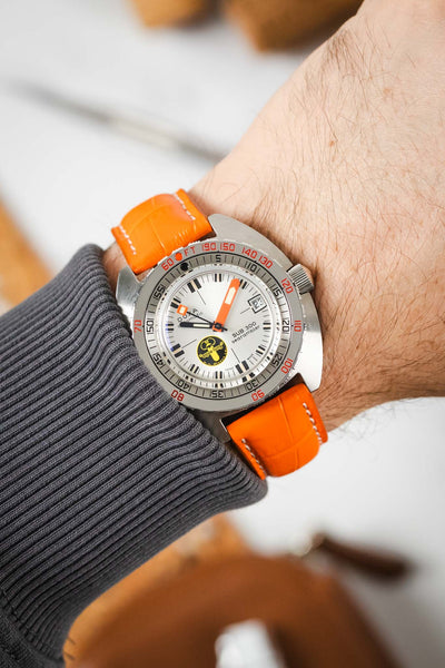 
A close-up shot of a person's wrist wearing a DOXA SUB 300 watch with a silver dial, orange accents, and an orange crocodile-embossed leather strap. The person is wearing a gray long-sleeve shirt. The background is softly blurred.