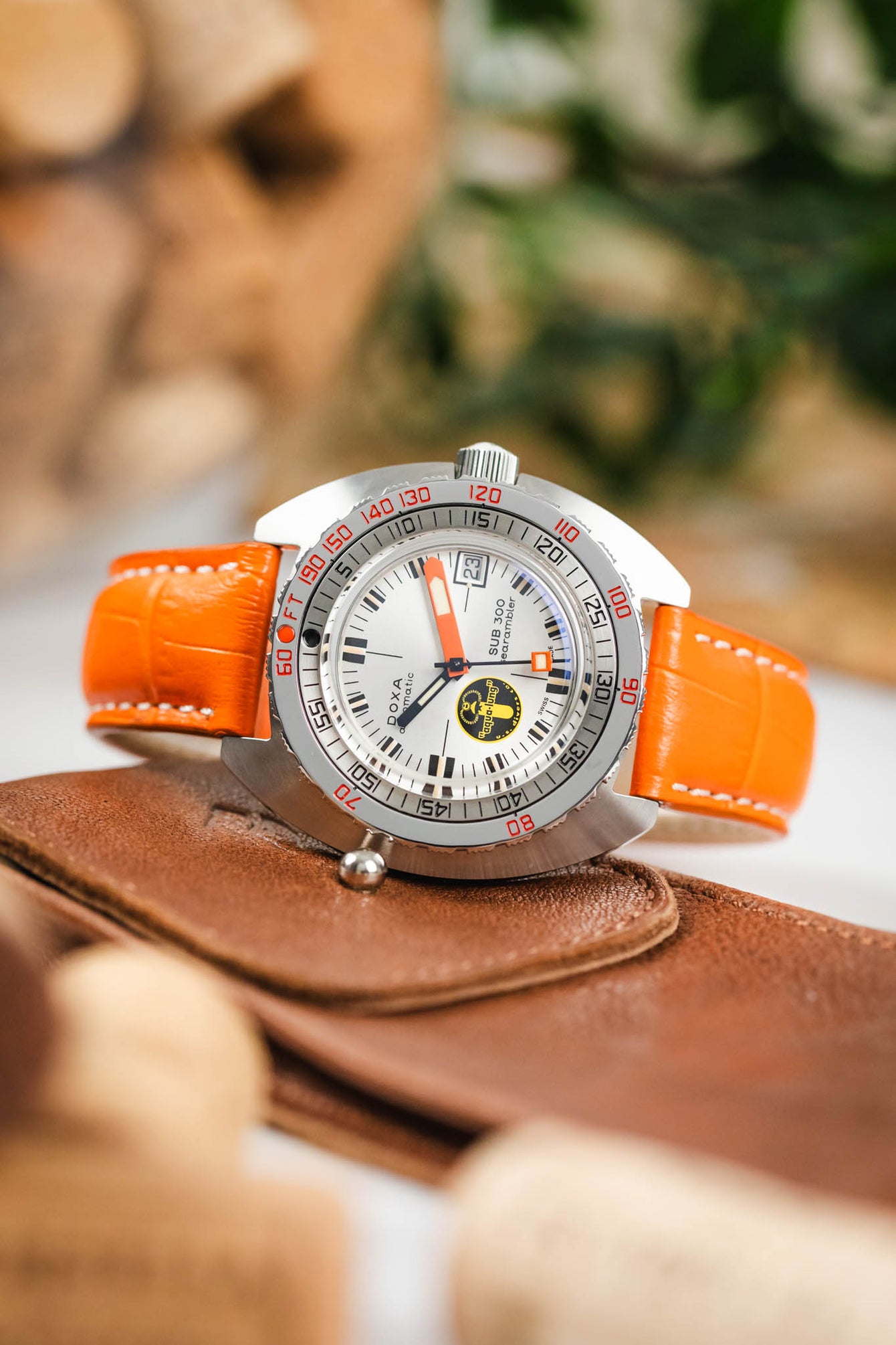 
A slightly elevated shot of a DOXA SUB 300 watch with a silver dial and an orange, crocodile-embossed leather strap. The watch is positioned on a tan leather watch pouch with a button closure. The background is softly blurred, showing corks and other elements.