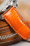 A close-up, macro shot focuses on a vibrant orange, crocodile-embossed leather watch strap with white contrast stitching. A silver watch case is visible at the top, while the strap rests on a brown leather watch pouch with a prominent zipper. The background is softly blurred.