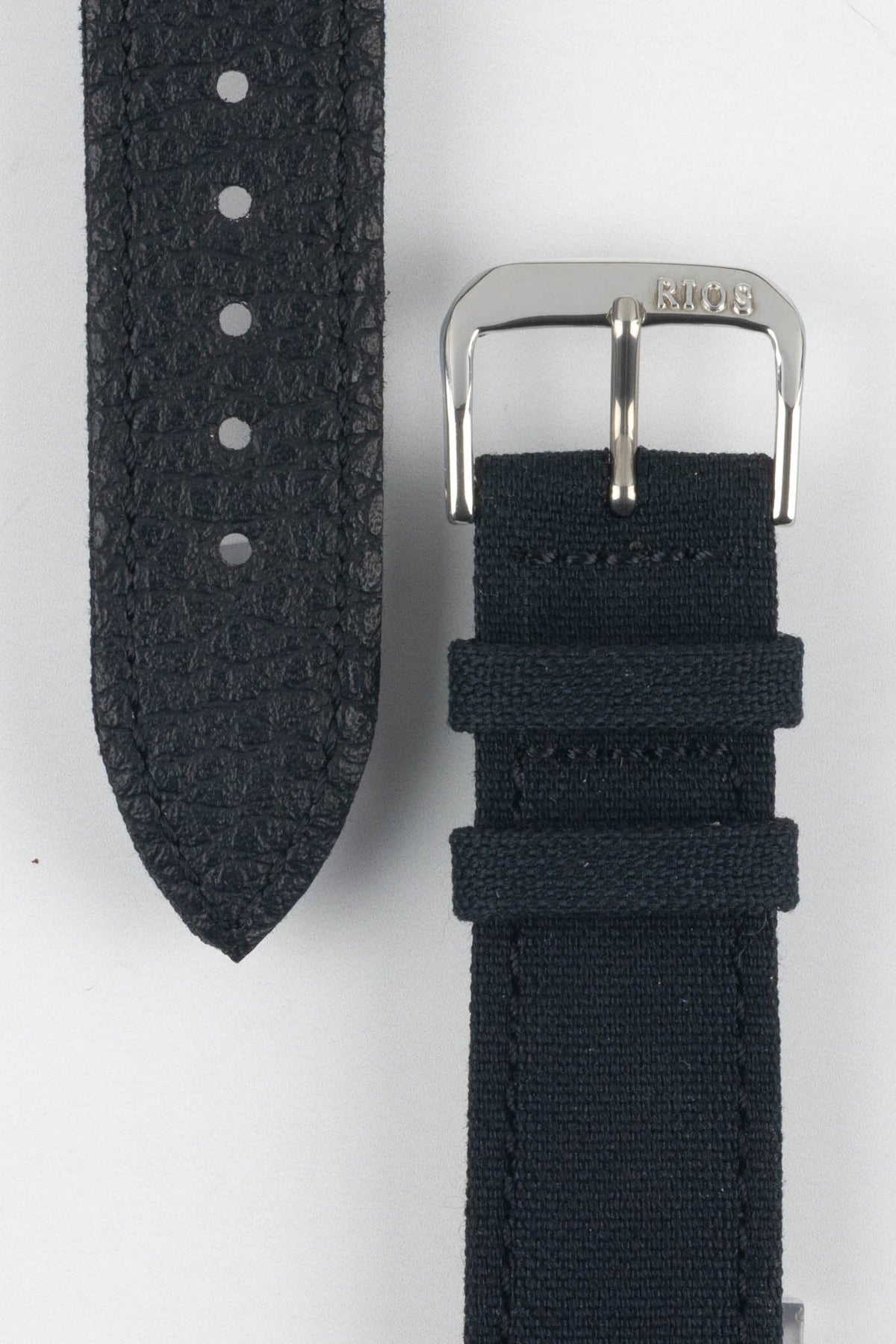 Close-up view of a dark navy watch strap showing both sides on a light gray background. The textured leather underside with adjustment holes and pointed tip is displayed alongside a woven fabric upper section featuring a polished silver-tone RIOS-branded buckle and two matching fabric keepers. The image highlights the strap’s contrasting materials, stitching, and construction details.