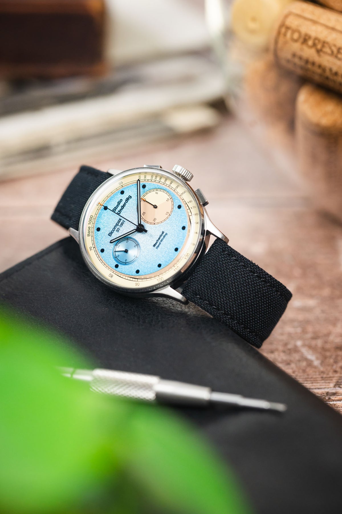 Studio Underd0g Bicompax chronograph watch on a black canvas strap, displayed on a leather surface with watchmaking tools and vintage accessories.
