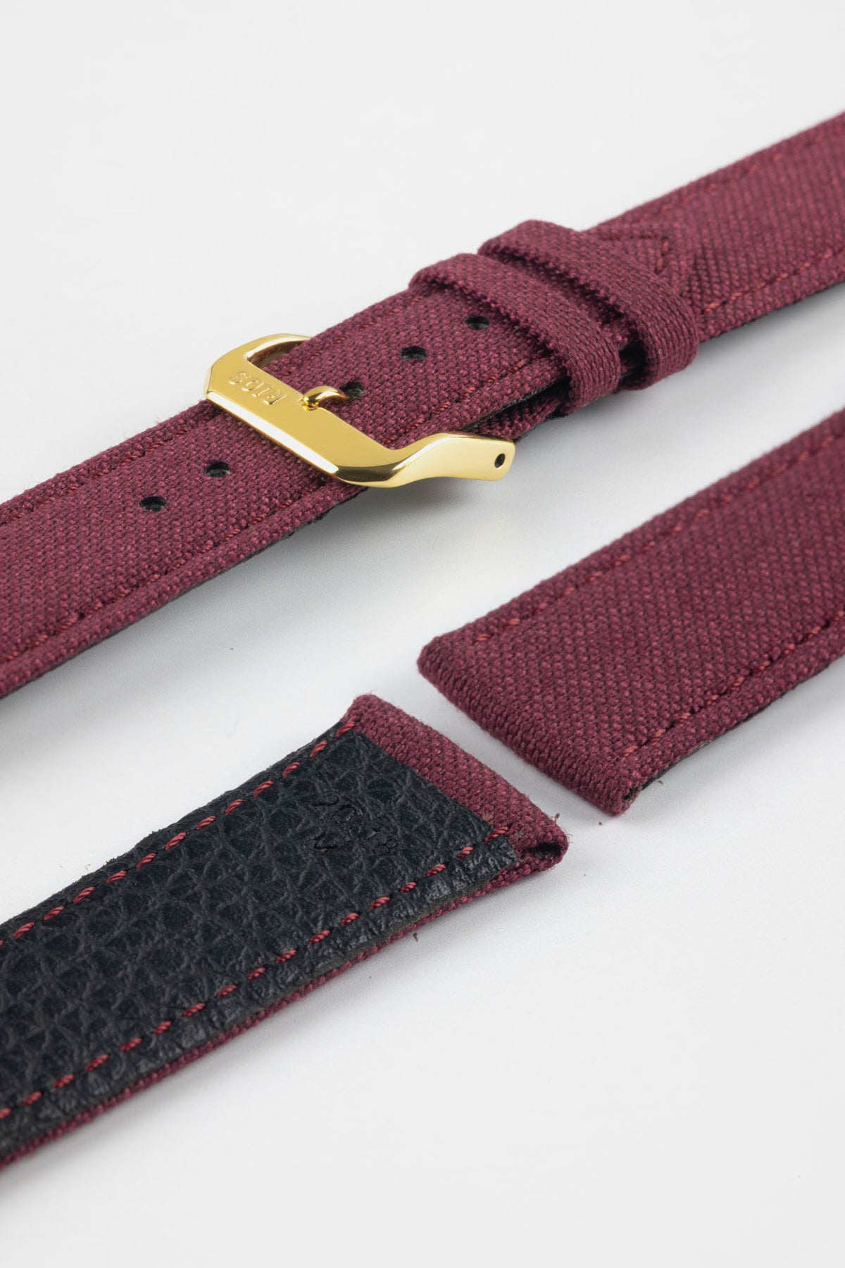 Close-up product image of a burgundy fabric watch strap displayed in separate sections on a light gray background. The strap features a polished gold-tone RIOS-branded buckle, matching fabric keepers, and stitched edges. A section of the black textured leather underside with burgundy contrast stitching is visible, highlighting the combination of woven fabric and leather backing, along with the strap’s premium construction and detailing.