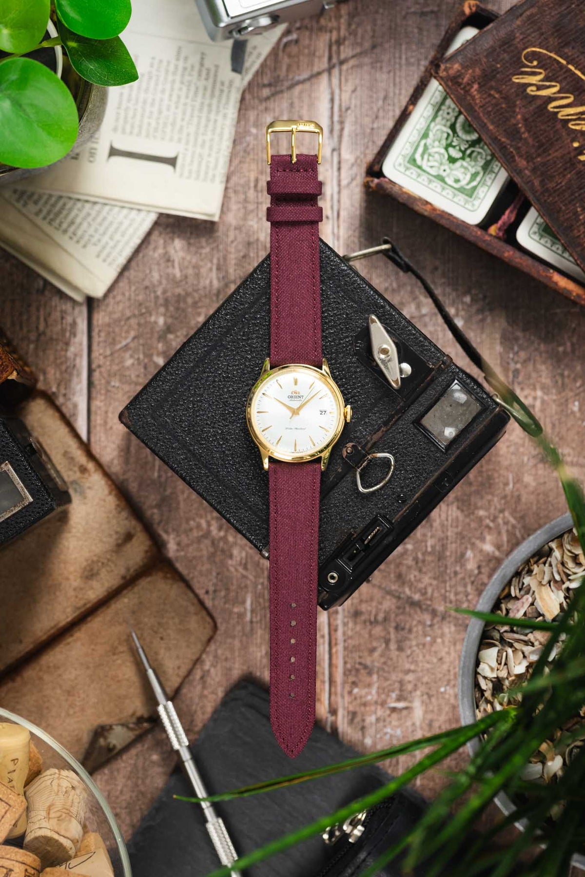 Orient Bambino automatic watch on a burgundy canvas strap, displayed with vintage accessories on a rustic wooden surface.