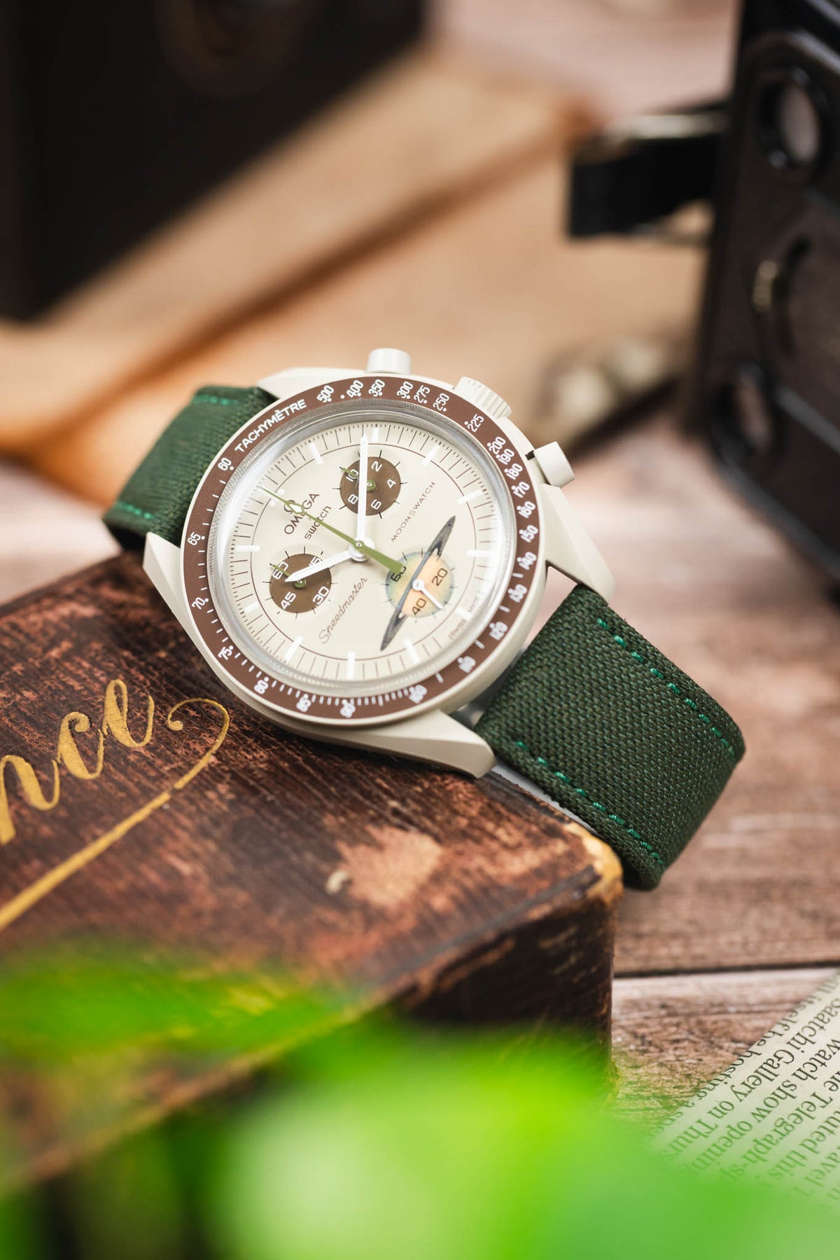 Omega x Swatch MoonSwatch Mission to Jupiter on an olive green canvas strap, displayed on a vintage wooden box with retro camera accessories.