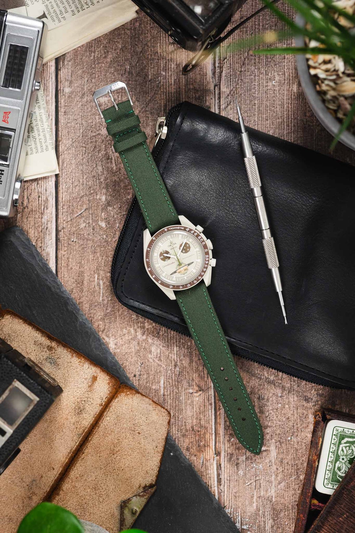 Omega x Swatch MoonSwatch Mission to Jupiter on an olive green canvas strap, displayed with vintage accessories and watchmaking tools on a rustic wooden surface.