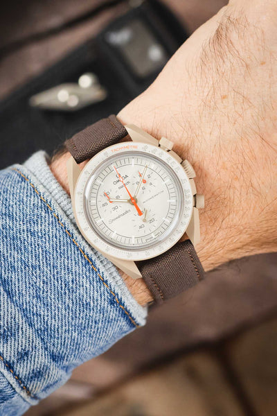 Omega x Swatch MoonSwatch on a mocha brown canvas strap, shown on wrist with a white chronograph dial and beige case.
