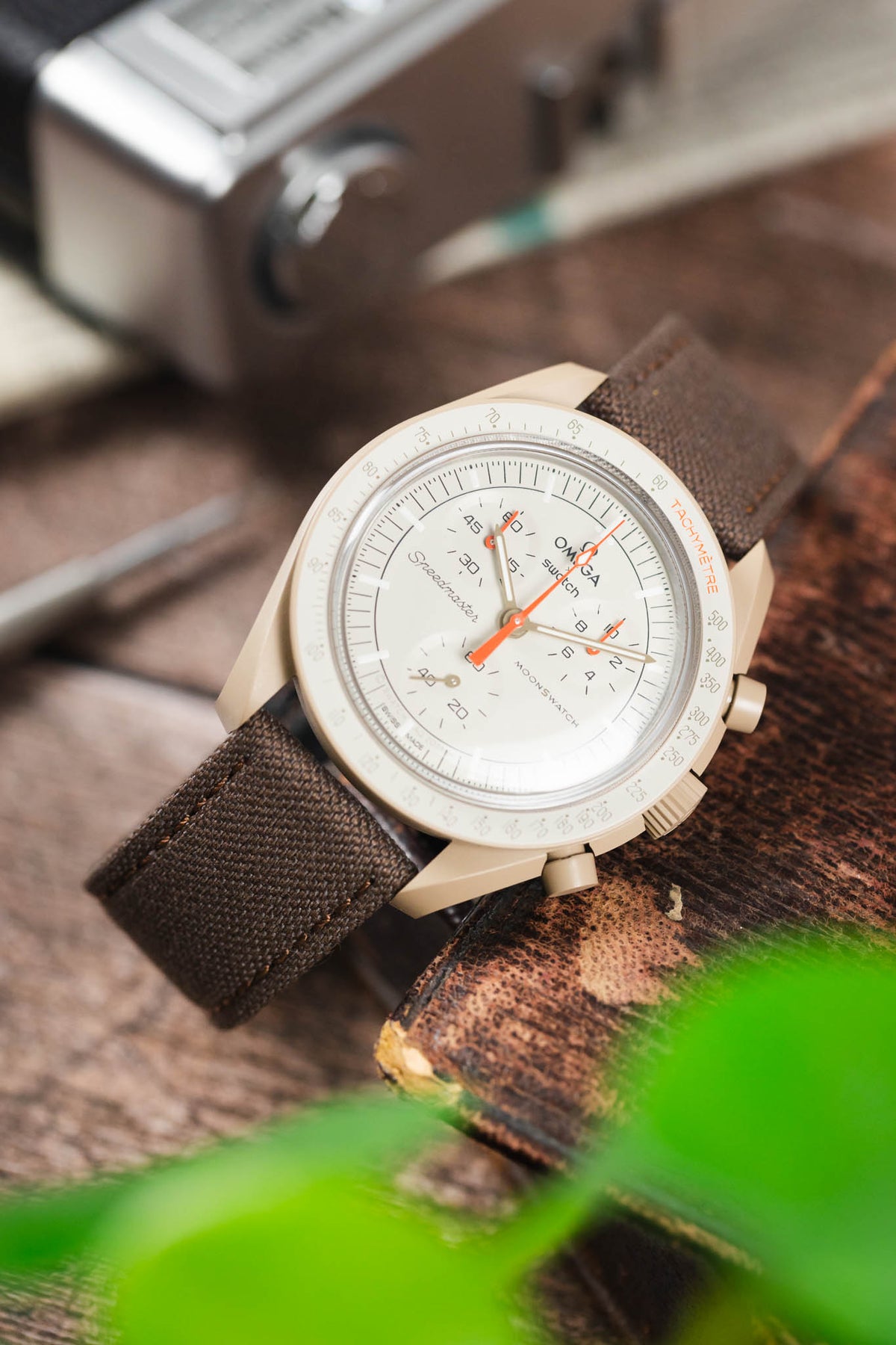 Omega x Swatch MoonSwatch on a mocha brown canvas strap, displayed on a vintage book with a retro camera and watchmaking tools.