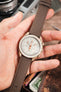 Omega x Swatch MoonSwatch on a mocha brown canvas strap, held in hand with a white chronograph dial and beige case.