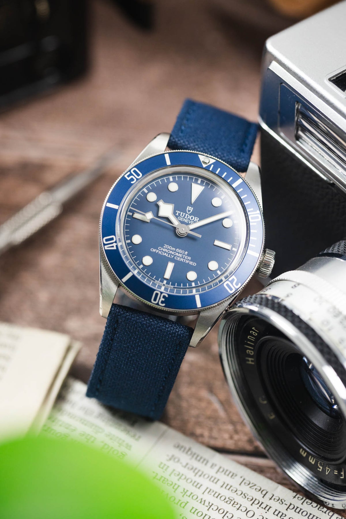 Tudor Black Bay dive watch on an ocean blue canvas strap, displayed beside a vintage camera on a rustic wooden surface.