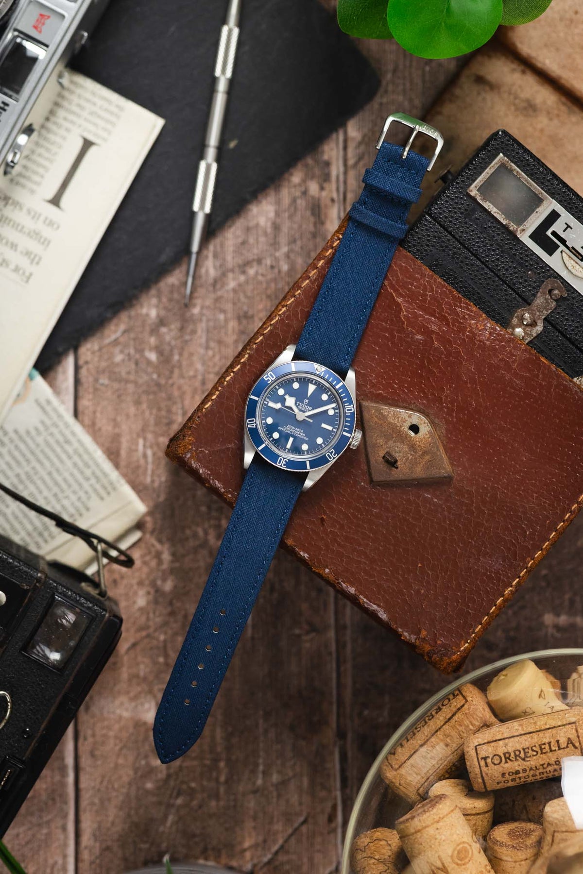 Tudor Black Bay dive watch on an ocean blue canvas strap, displayed with vintage accessories on a rustic wooden surface.
