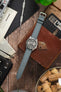 Seiko 5 automatic watch on a stone grey canvas strap, displayed with vintage accessories on a rustic wooden surface.
