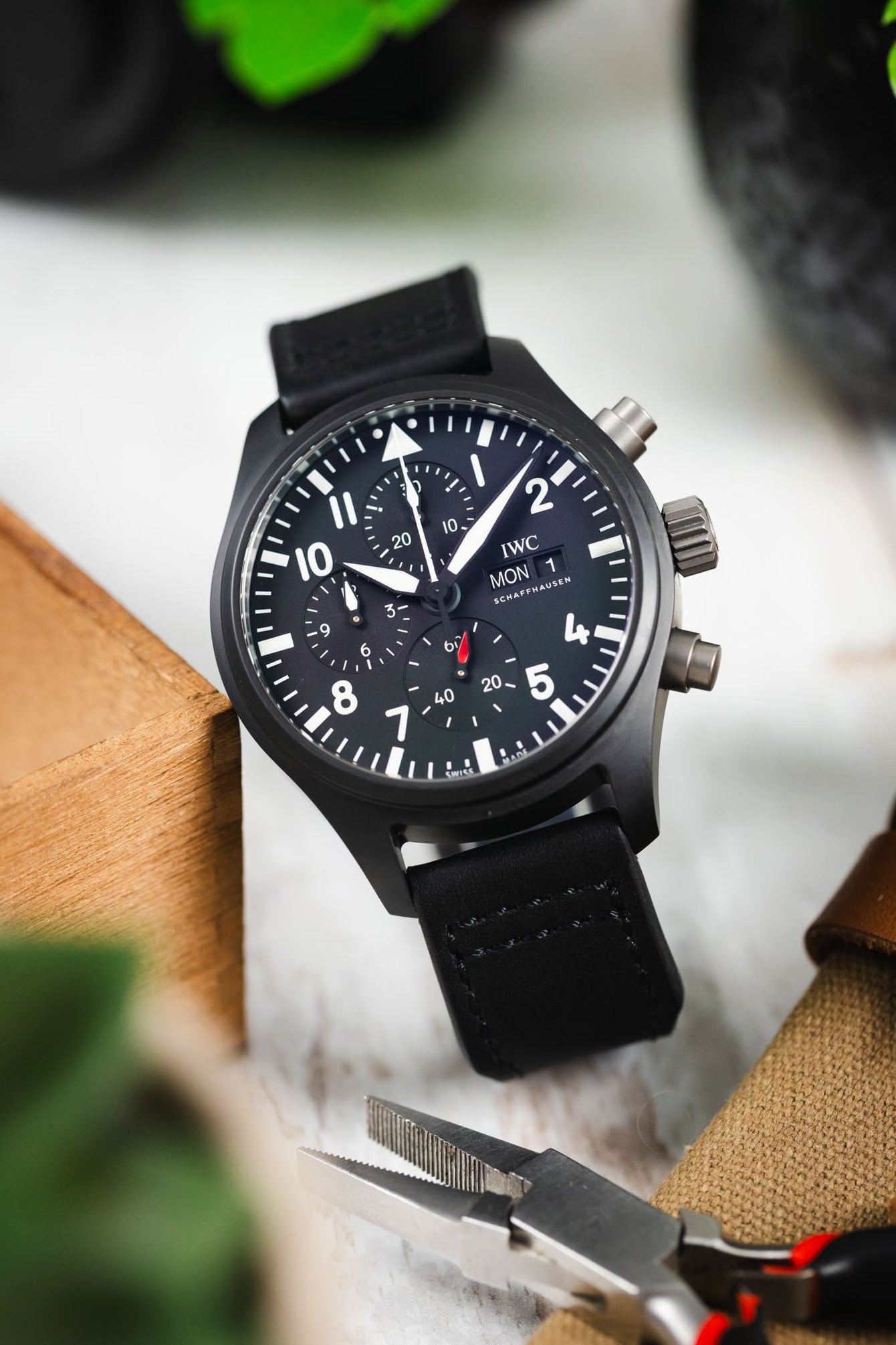 Close-up of a black IWC chronograph wristwatch with a black leather strap placed at an angle on a light surface, surrounded by pliers, fabric, and blurred green leaves, highlighting the dark dial with white numerals and subdials.