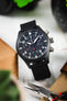 Close-up of a black IWC chronograph wristwatch with a black leather strap placed at an angle on a light surface, surrounded by pliers, fabric, and blurred green leaves, highlighting the dark dial with white numerals and subdials.