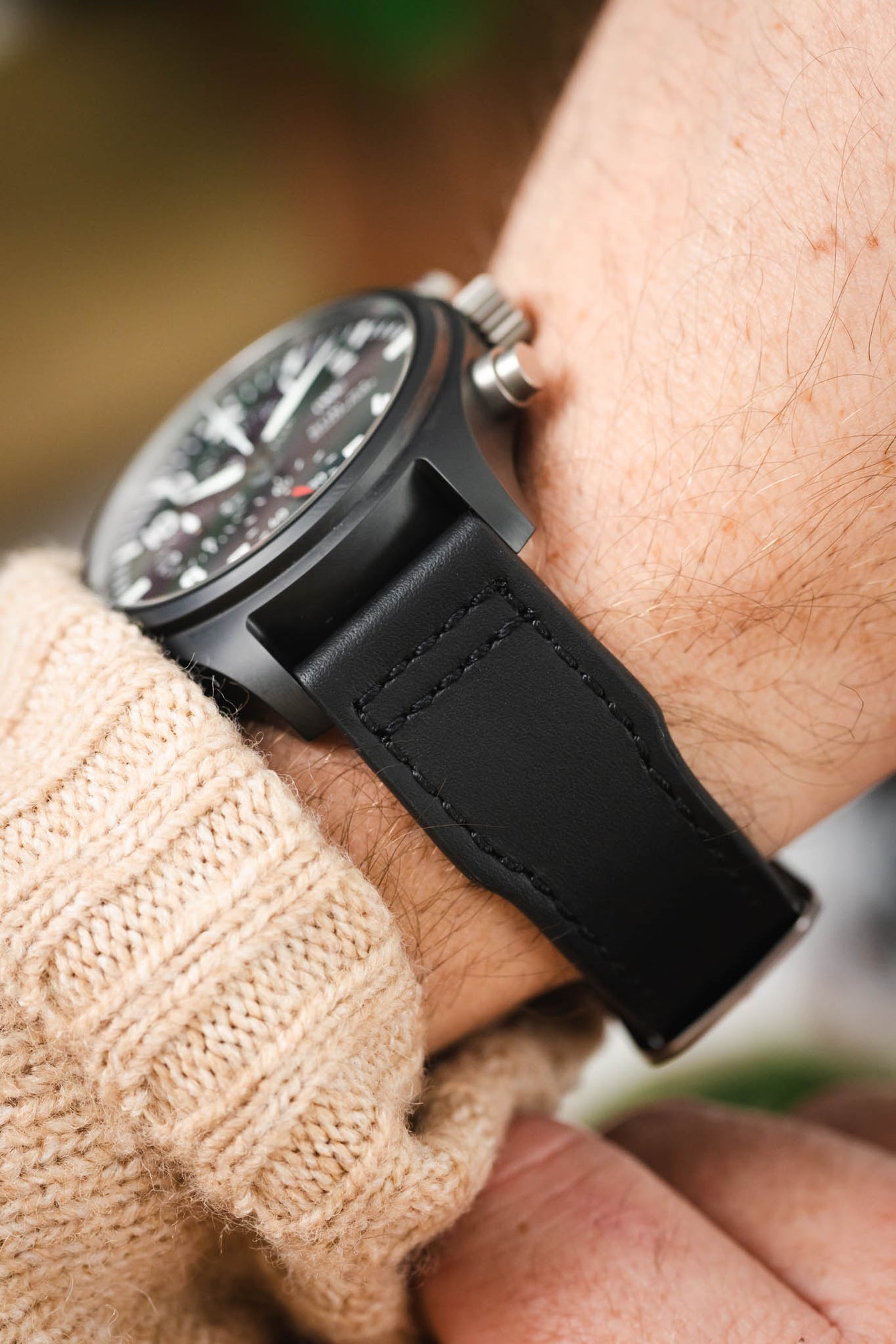 Macro side view of a black IWC chronograph wristwatch with a black leather strap worn on a wrist, highlighting the stitching detail, matte case, and pushers, with a beige knit sleeve and softly blurred background.