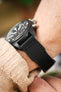 Macro side view of a black IWC chronograph wristwatch with a black leather strap worn on a wrist, highlighting the stitching detail, matte case, and pushers, with a beige knit sleeve and softly blurred background.