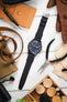 Flat lay of a black IWC chronograph wristwatch with a black leather strap centered on a light wooden surface, surrounded by a tan leather pouch, brown gloves, pliers, a watch tool, canvas fabric, a leather strap, and a wooden box with checkers and dice pieces.