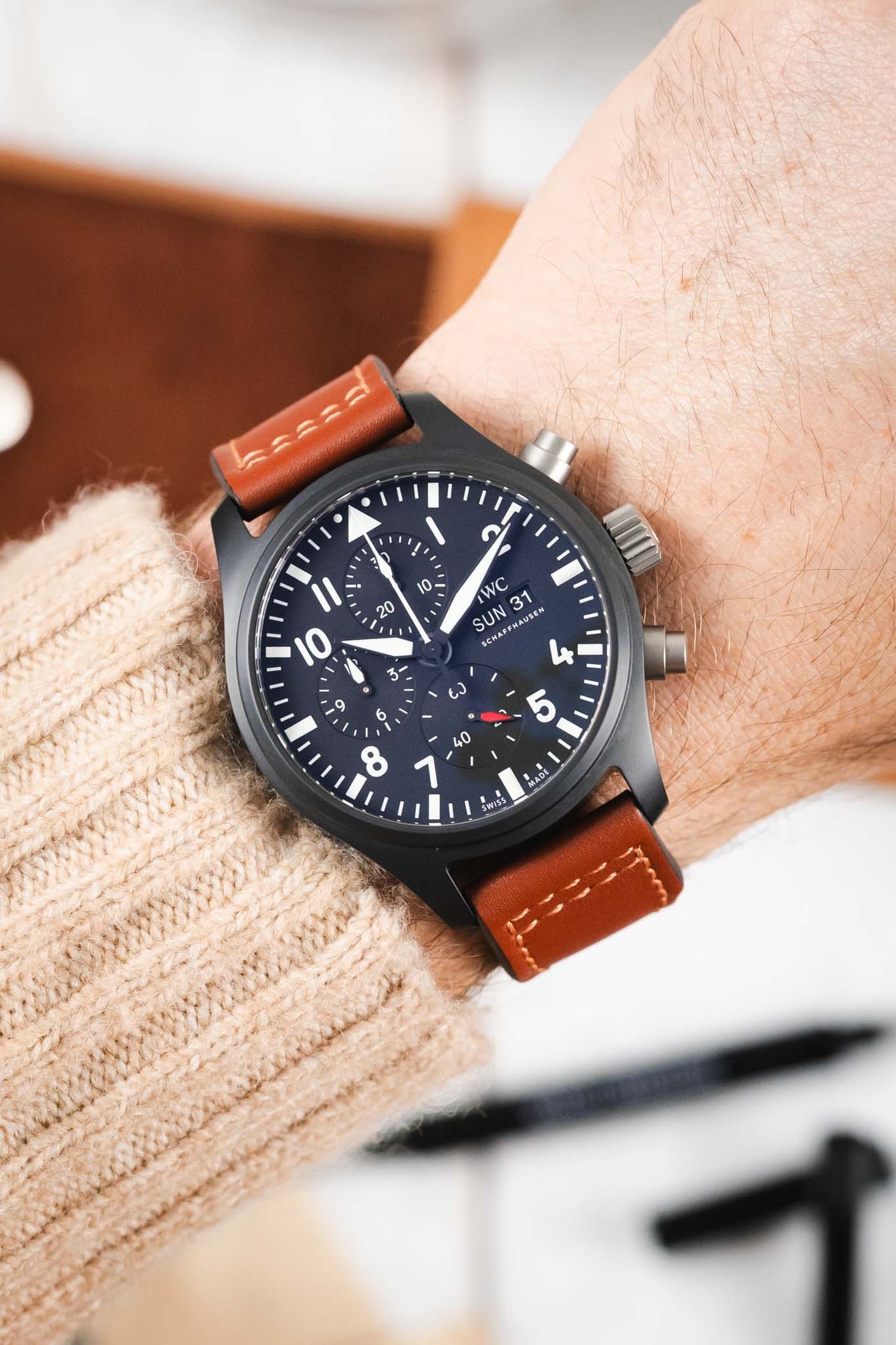 Close-up of a black IWC chronograph wristwatch with a brown leather strap worn on a wrist, featuring a dark dial with white numerals and subdials, paired with a beige knit sleeve in a softly lit indoor setting.