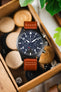 Close-up of a black IWC chronograph wristwatch with a brown leather strap placed over an open wooden box containing round wooden discs and small dice, with green plant leaves in the background.