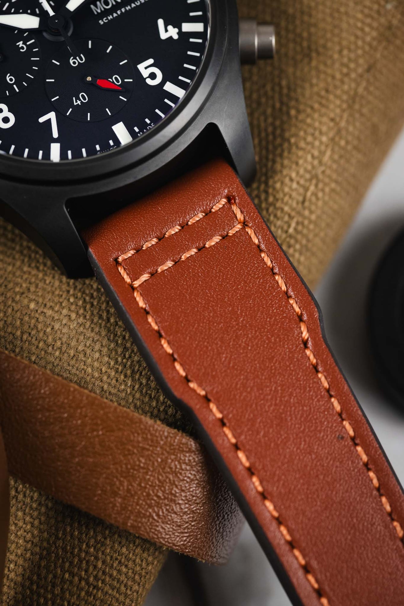 Macro close-up of a black IWC chronograph watch case and brown leather strap, highlighting detailed stitching and textured leather grain against a fabric background.