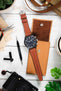 Flat lay of a black IWC chronograph wristwatch with a brown leather strap placed on a tan leather pouch, surrounded by watch tools, a camera, fabric, and small accessories on a light wooden surface with green plant accents.