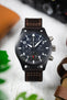 Close-up product shot of a black IWC chronograph watch with a dark dial, white numerals, three subdials, and a day-date window, mounted on a brown leather strap. The watch is centered on a light surface, with blurred green leaves and leather accessories in the background.