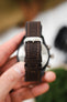 Close-up of a brown leather watch strap with contrast stitching and a polished metal buckle engraved “RIOS,” held in front of a blurred watch face. The strap shows adjustment holes and keeper loops, with a softly blurred background featuring green foliage and leather items.