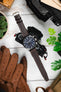 Flat lay of a brown leather watch strap attached to a black IWC chronograph wristwatch resting on a camera and lens, surrounded by brown leather driving gloves, green potted plants, and part of a hat on a light wooden surface.