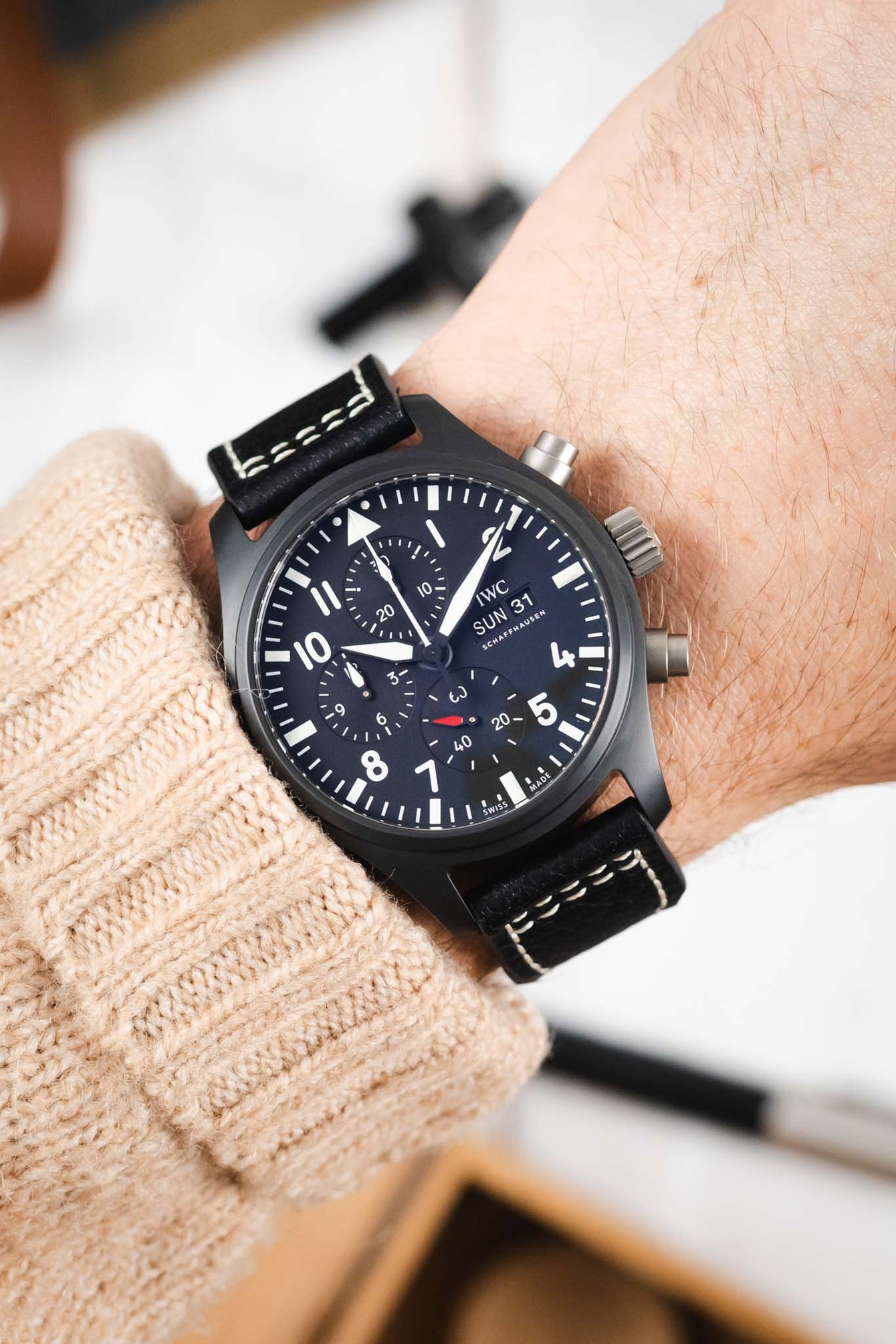 Close-up of a black IWC chronograph wristwatch with a black leather strap and white contrast stitching, worn on a wrist with a beige knit sleeve, showing a dark dial with bold white numerals, subdials, and date display against a softly blurred indoor background.