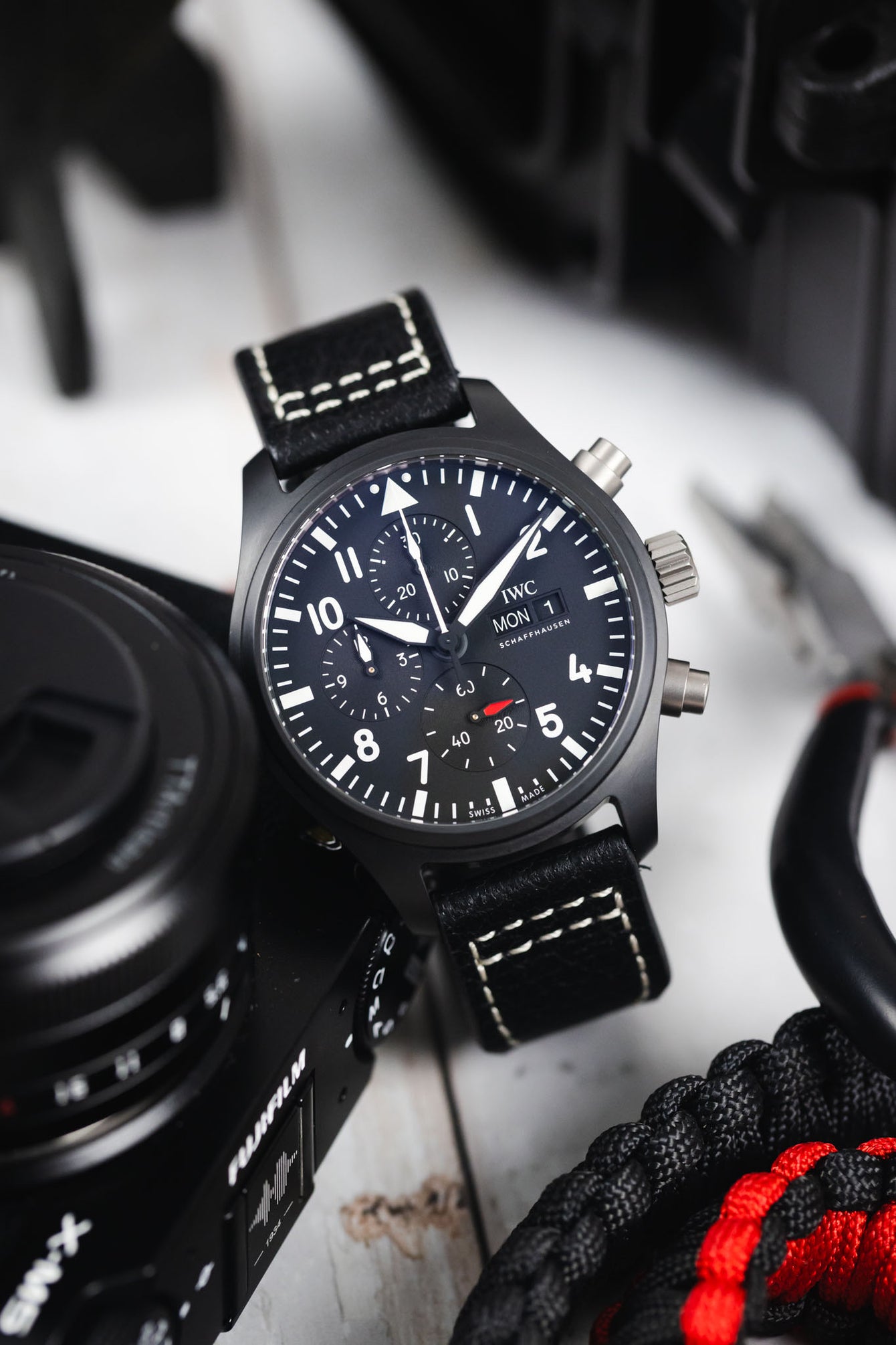 Close-up of a black IWC chronograph wristwatch with a black leather strap and white contrast stitching resting against a camera, with paracord rope and tools nearby, highlighting the dark dial with bold white numerals, subdials, and date display on a light surface.
