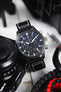 Close-up of a black IWC chronograph wristwatch with a black leather strap and white contrast stitching resting against a camera, with paracord rope and tools nearby, highlighting the dark dial with bold white numerals, subdials, and date display on a light surface.