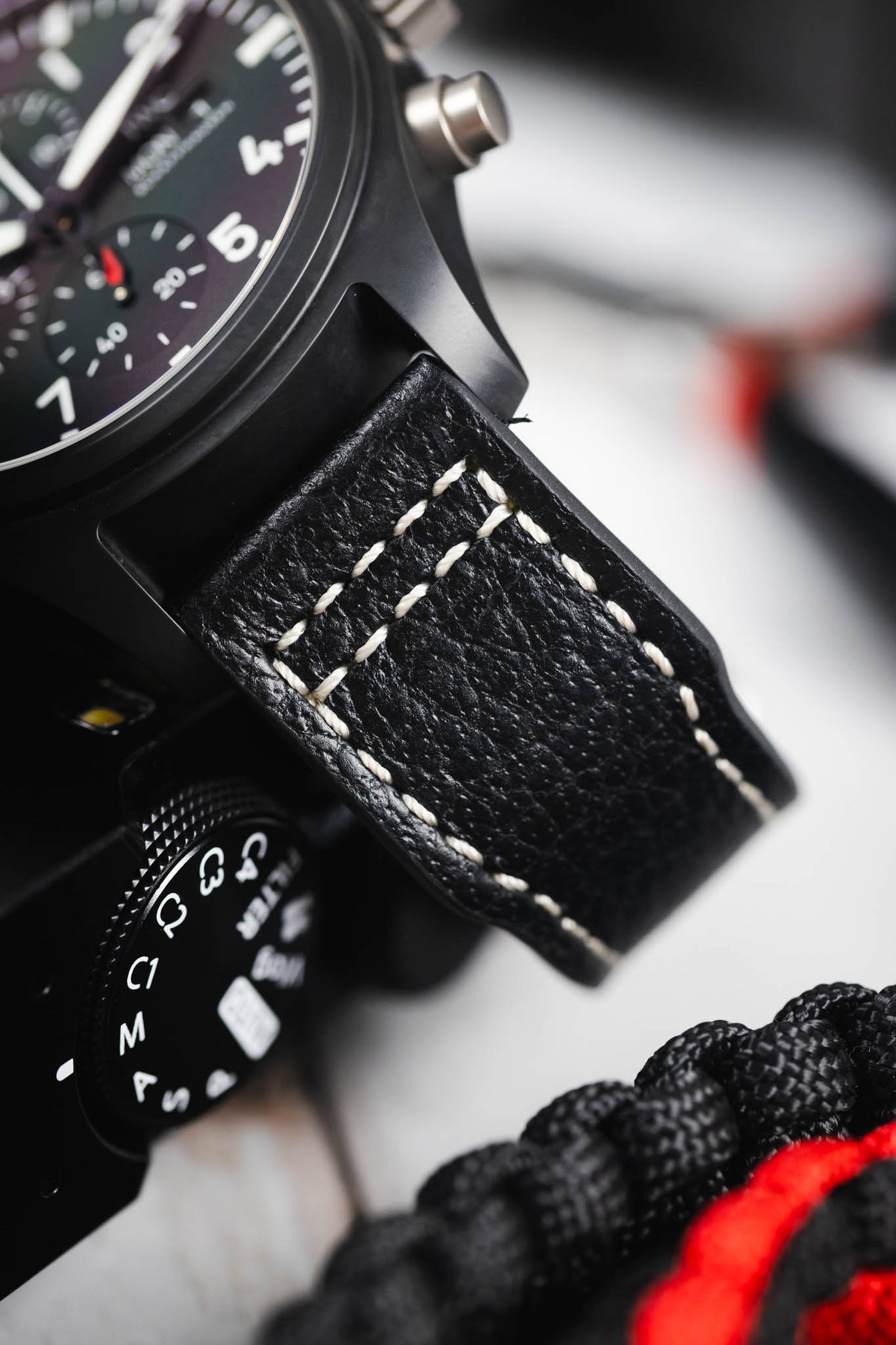 Macro close-up of a black IWC chronograph watch with a textured black leather strap featuring white contrast stitching, showing the matte case, pushers, and part of the dial, positioned next to a camera and braided cord on a light surface.