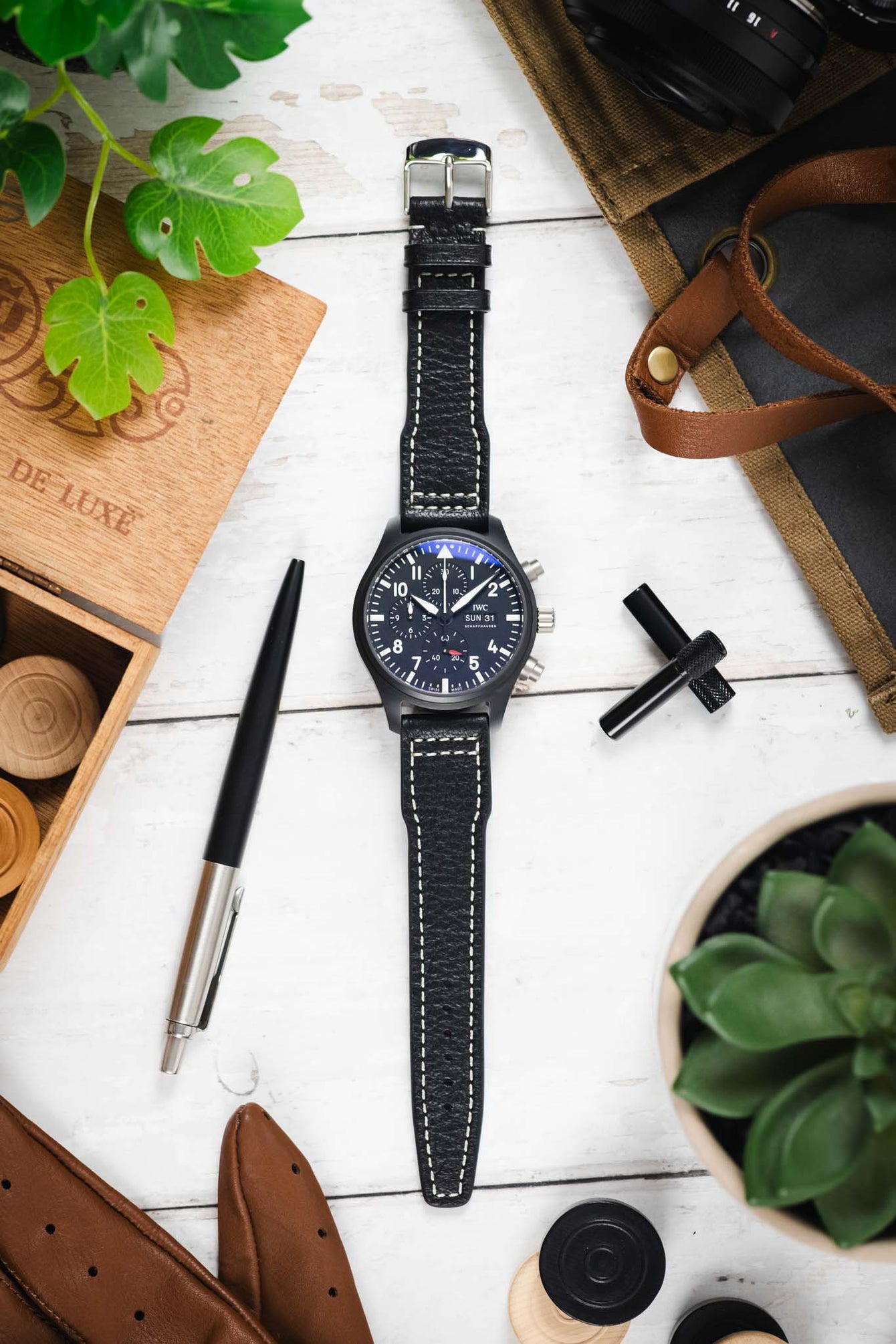 Flat lay of a black IWC chronograph wristwatch with a textured black leather strap and white contrast stitching, centered on a light wooden surface and surrounded by a wooden box with game pieces, leather gloves, a pen, a watch tool, canvas fabric, a bag strap, and green potted plants.