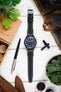 Flat lay of a black IWC chronograph wristwatch with a textured black leather strap and white contrast stitching, centered on a light wooden surface and surrounded by a wooden box with game pieces, leather gloves, a pen, a watch tool, canvas fabric, a bag strap, and green potted plants.