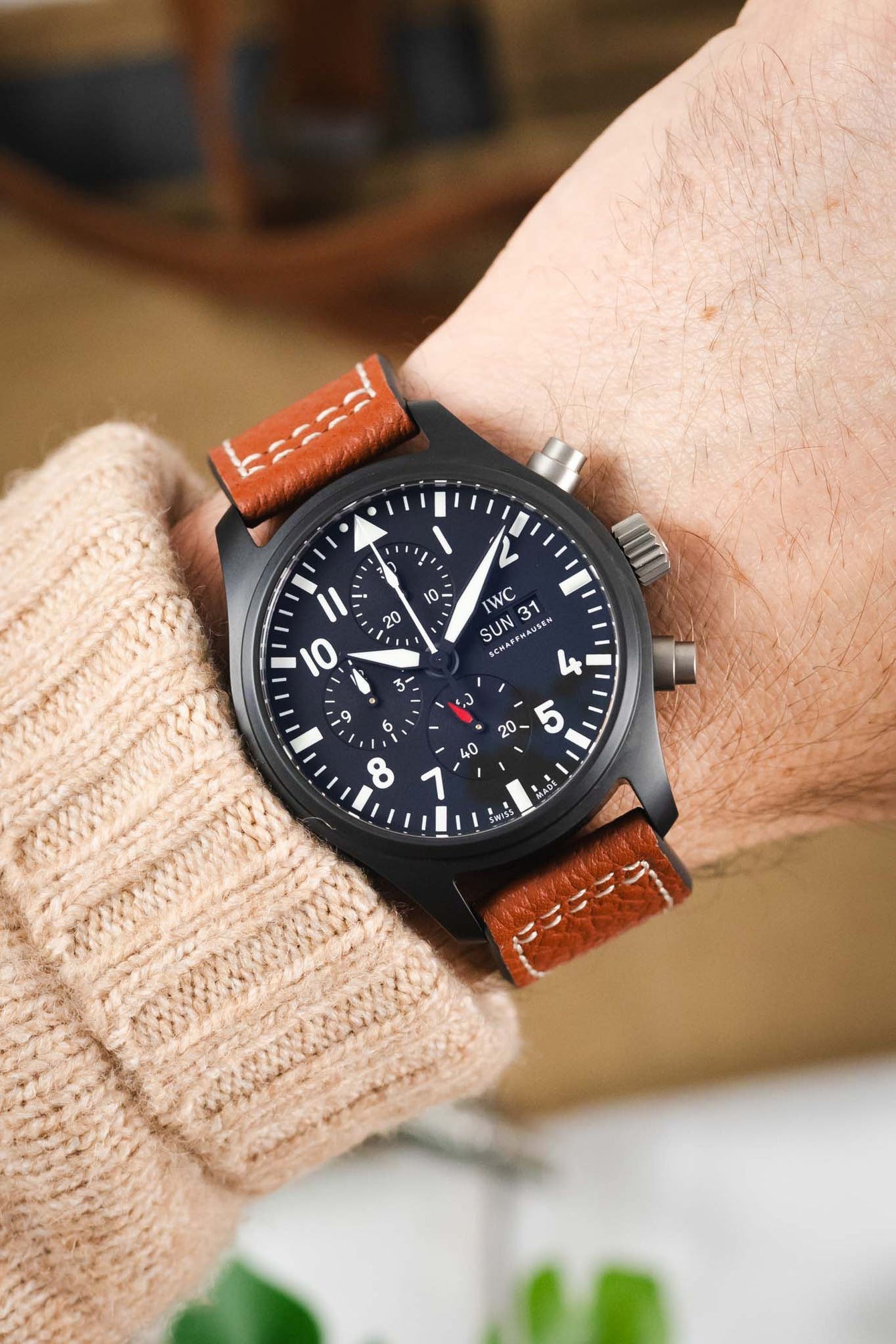Close-up of a black IWC chronograph wristwatch with a dark dial, bold white numerals, subdials, and a day-date display, worn on a wrist with a cognac-brown leather strap featuring white contrast stitching, paired with a beige knit sleeve and set against a softly blurred warm-toned background.