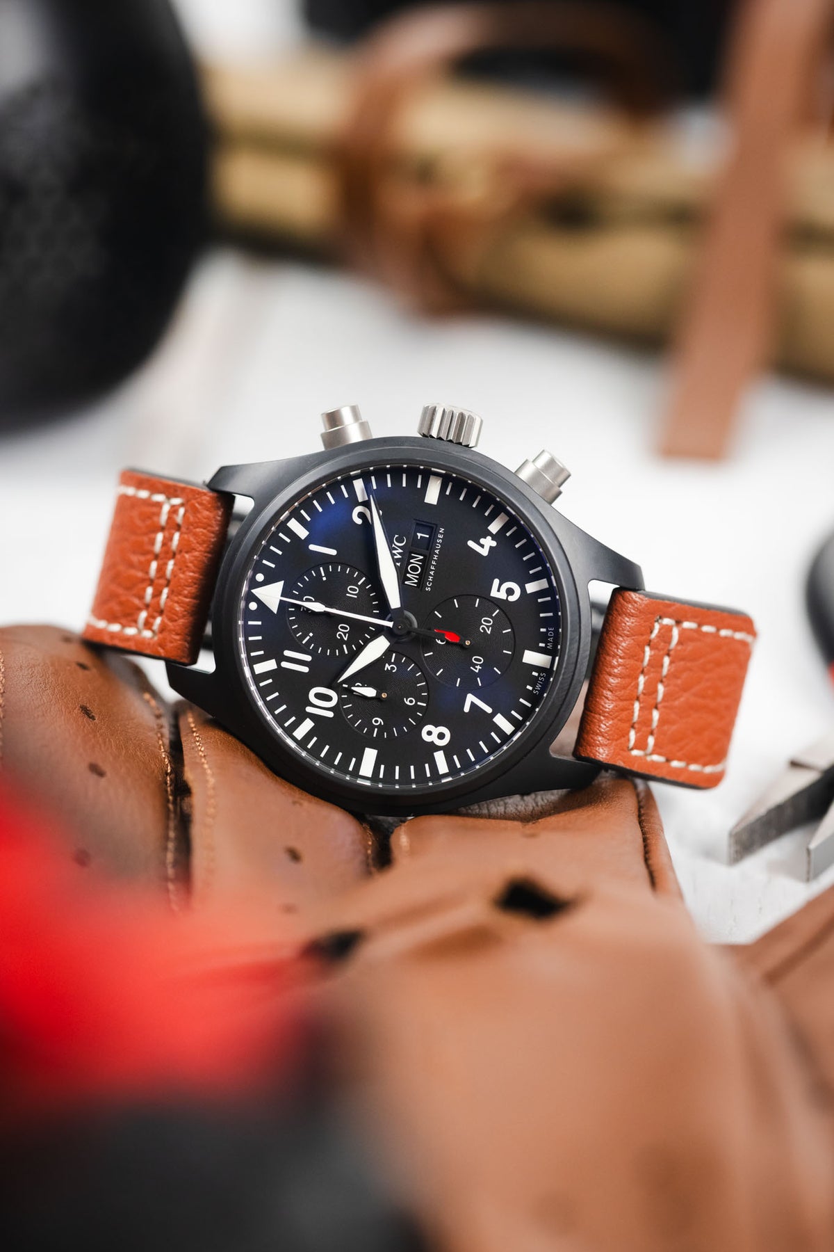 Close-up of a black IWC chronograph wristwatch with a matte case, dark dial, bold white numerals, three subdials, and a day-date display, paired with a cognac-brown leather strap with white contrast stitching, resting on brown leather gloves with a blurred background featuring tools and warm-toned objects.