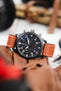 Close-up of a black IWC chronograph wristwatch with a matte case, dark dial, bold white numerals, three subdials, and a day-date display, paired with a cognac-brown leather strap with white contrast stitching, resting on brown leather gloves with a blurred background featuring tools and warm-toned objects.