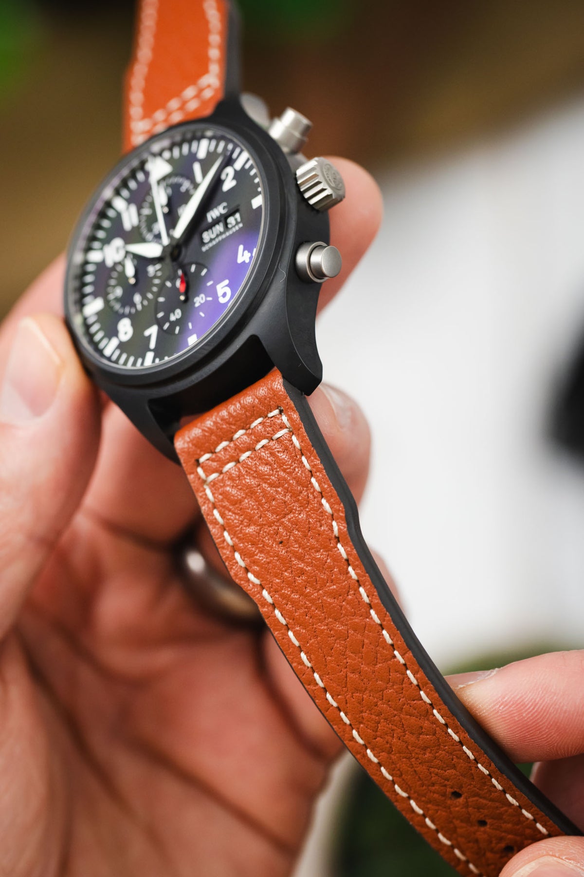 Macro close-up of a black IWC chronograph wristwatch with a matte case, showing the side profile with crown and pushers, paired with a cognac-brown leather strap featuring white contrast stitching, held in hand against a softly blurred background with warm tones and greenery.