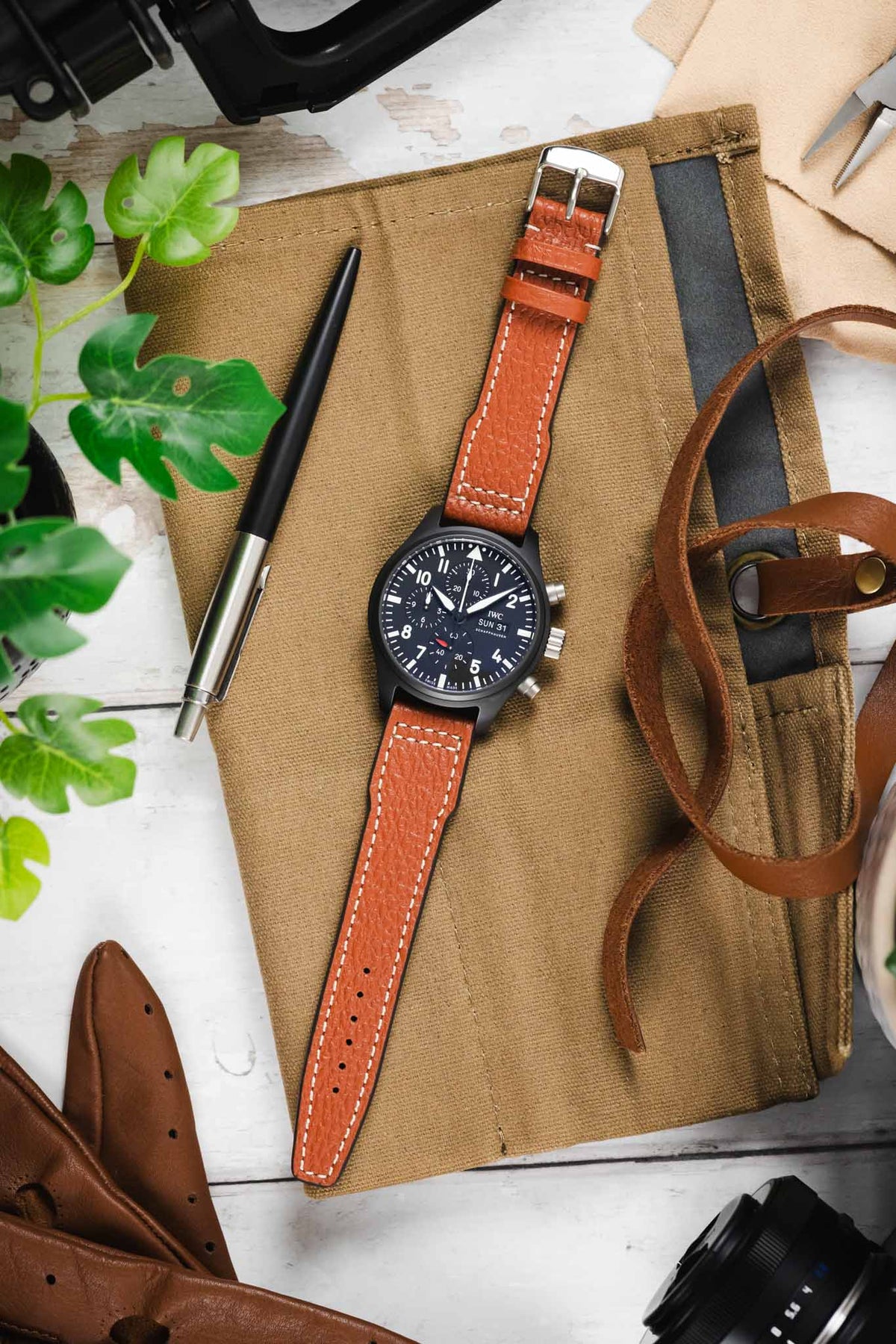Flat lay of a black IWC chronograph wristwatch with a dark dial, bold white numerals, subdials, and a day-date display, paired with a cognac-brown leather strap with white contrast stitching, positioned on a tan canvas roll alongside a pen, leather strap, small tools, green plant leaves, and camera gear on a light wooden surface.