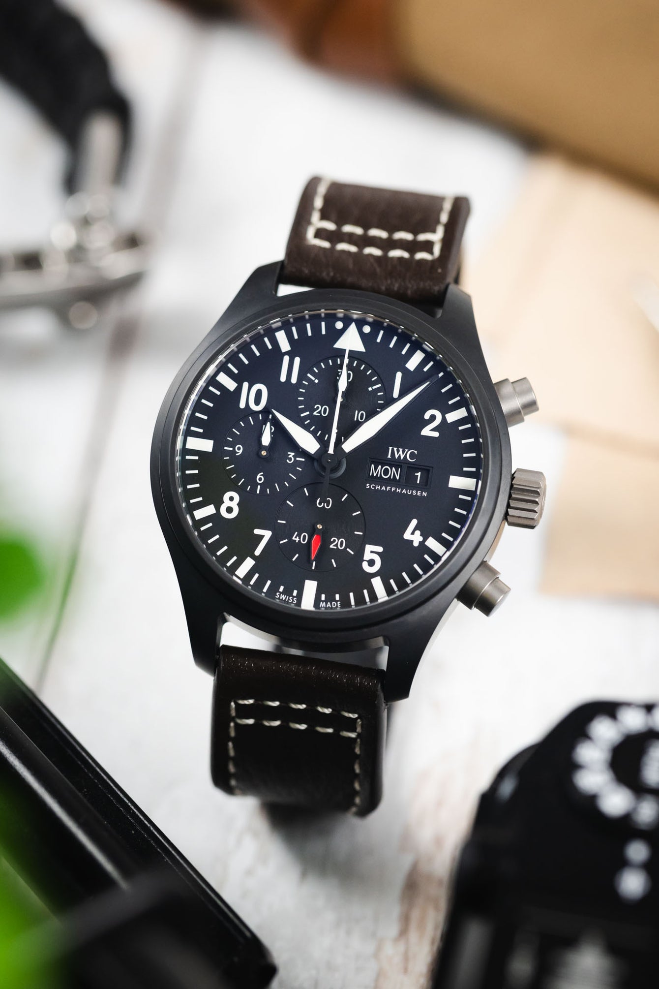 Close-up of a black IWC chronograph wristwatch with a dark dial, bold white numerals, subdials, and a day-date display, paired with a brown leather strap featuring white contrast stitching, positioned upright on a light wooden surface with a blurred background including tools, a box, and greenery.