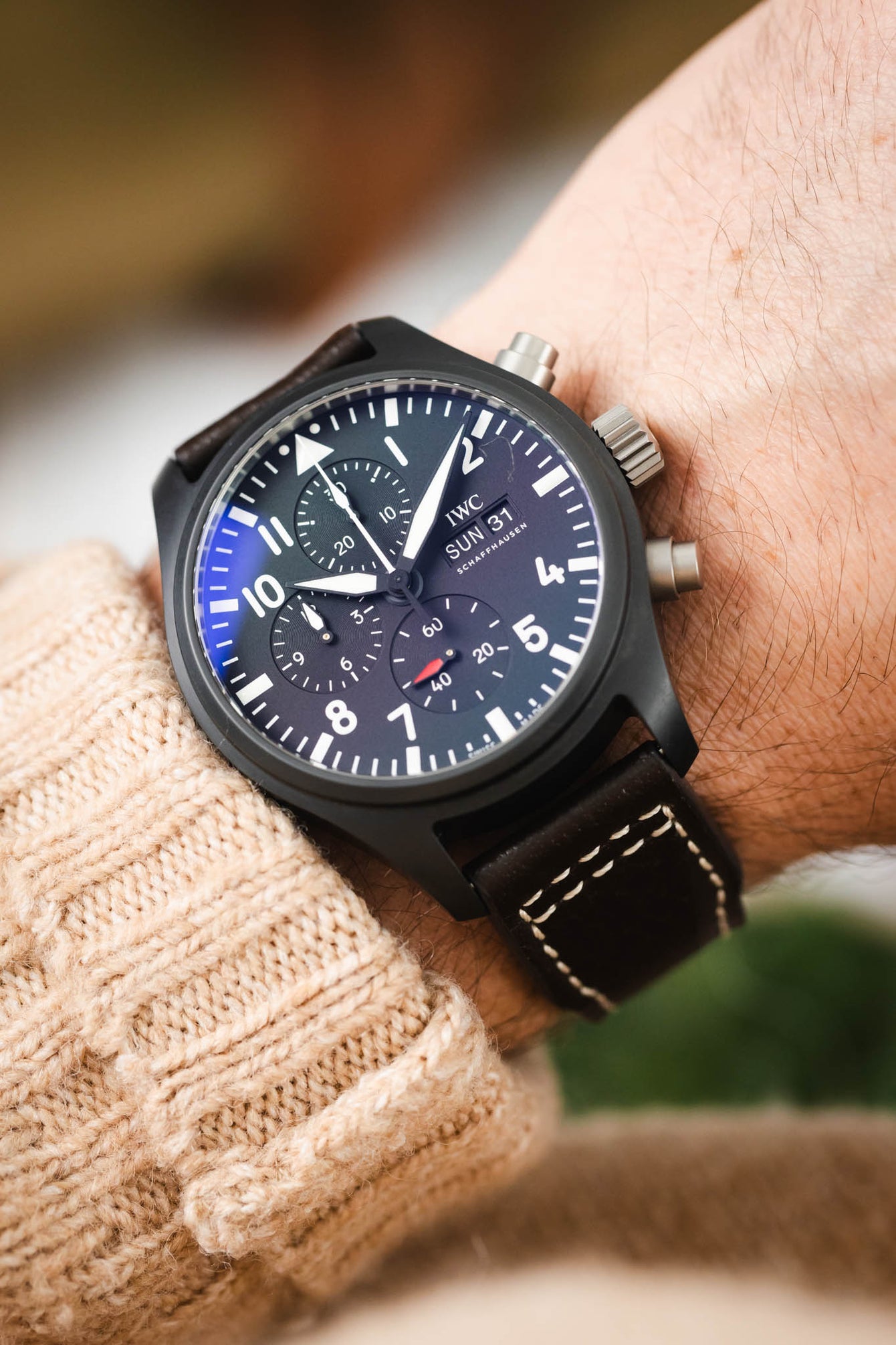 Close-up of a black IWC chronograph wristwatch with a dark dial, bold white numerals, subdials, and a day-date display, worn on a wrist with a brown leather strap featuring white contrast stitching, paired with a beige knit sleeve and set against a softly blurred background with warm tones and green plant leaves.