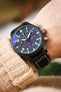 Close-up of a black IWC chronograph wristwatch with a dark dial, bold white numerals, subdials, and a day-date display, worn on a wrist with a brown leather strap featuring white contrast stitching, paired with a beige knit sleeve and set against a softly blurred background with warm tones and green plant leaves.
