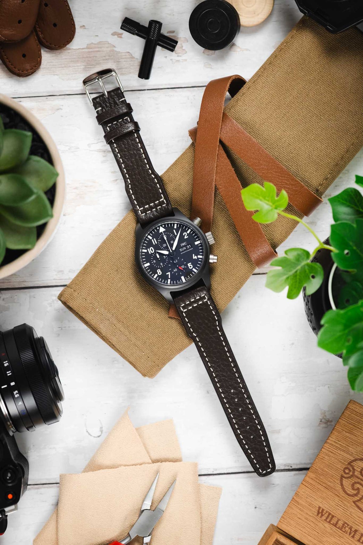 Flat lay of a black IWC chronograph wristwatch with a dark dial, bold white numerals, subdials, and day-date display, paired with a brown leather strap with white contrast stitching, placed on a tan canvas pouch with a leather strap, and surrounded by a camera, watch tools, cloth, wooden box, and green potted plants on a light wooden surface.
