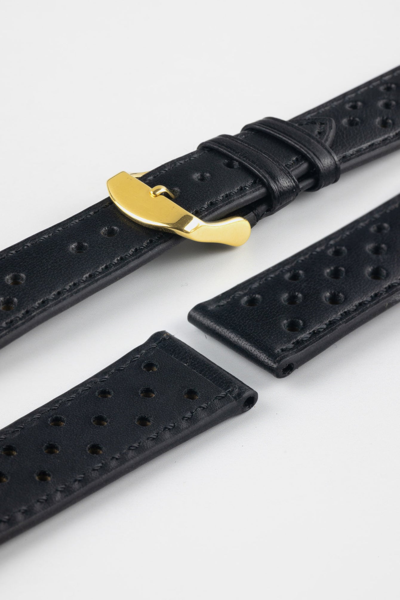 Close-up product shot of black perforated leather watch strap components with matching black stitching, including one fitted with a polished gold-tone deployment clasp. Additional strap sections are arranged on a plain light background, highlighting the leather grain, perforations, stitching detail, and gold clasp hardware.