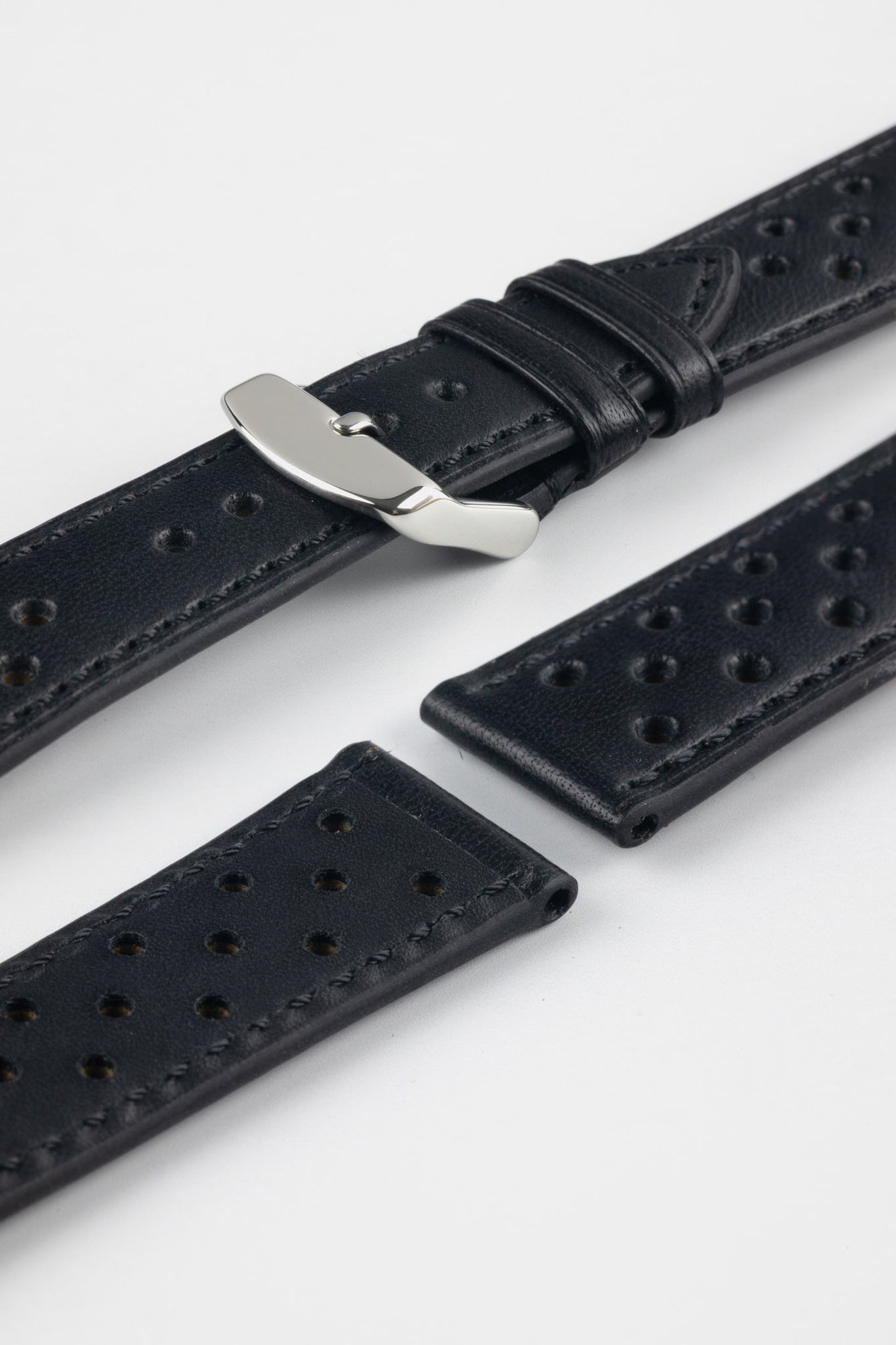 Close-up product shot of black perforated leather watch strap components with matching black stitching, including one fitted with a polished silver deployment clasp. Additional strap sections are arranged on a plain light background, highlighting the leather grain, perforations, stitching detail, and metal clasp hardware.