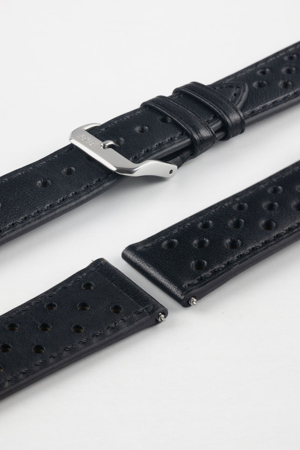 Close-up detail view of a black perforated leather rally watch strap displayed with one strap section detached, featuring black stitching, multiple motorsport-inspired perforations, dual keeper loops, a polished stainless steel RIOS buckle, quick-release spring bars, and a black leather lining. Displayed on a light grey background to highlight the premium handcrafted construction, tone-on-tone detailing, and vintage racing-inspired design.