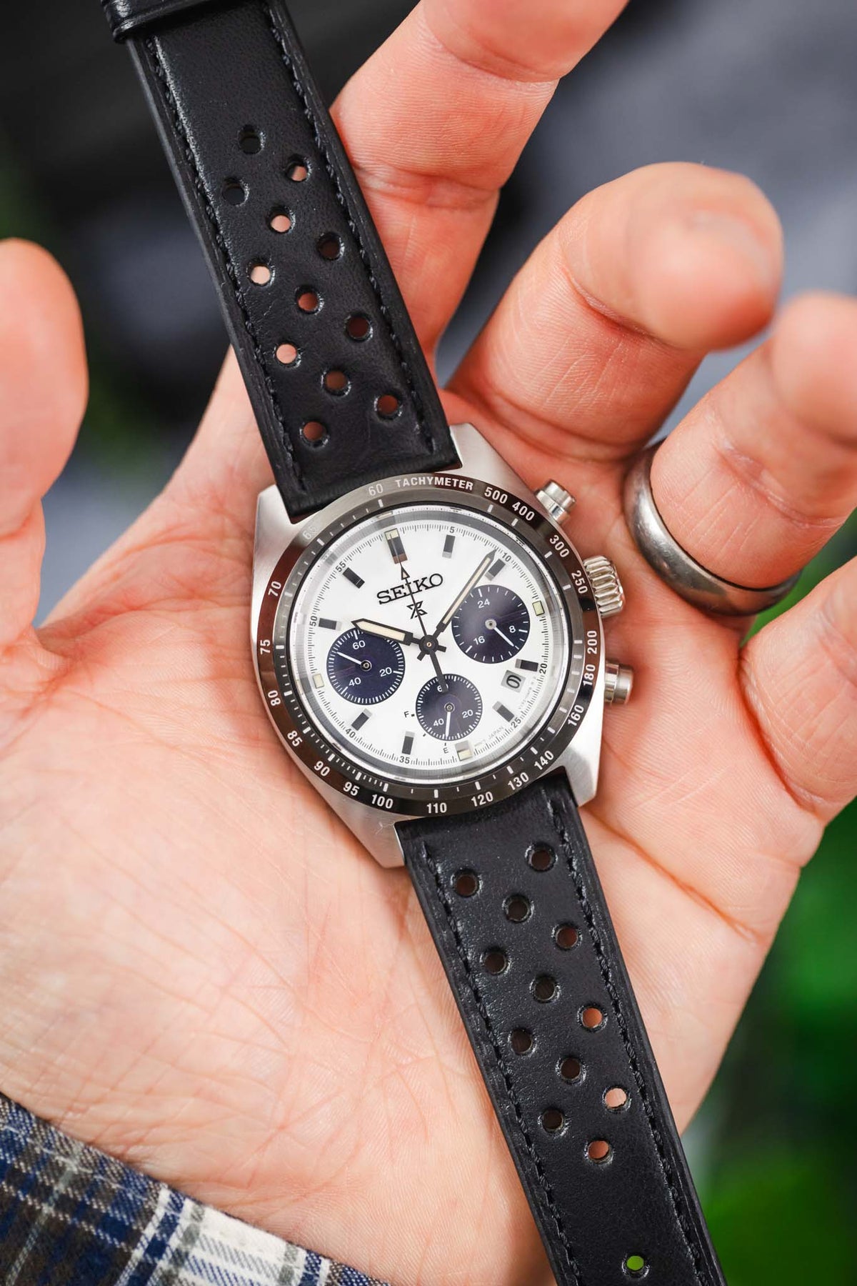 Close-up of a Seiko Speedtimer chronograph watch featuring a white panda dial with three dark blue sub-dials, black tachymeter bezel, and brushed stainless steel case, held in hand and fitted with a black perforated leather rally strap. The image highlights the strap's smooth leather finish, motorsport-inspired perforations, black stitching, and premium handcrafted construction.
