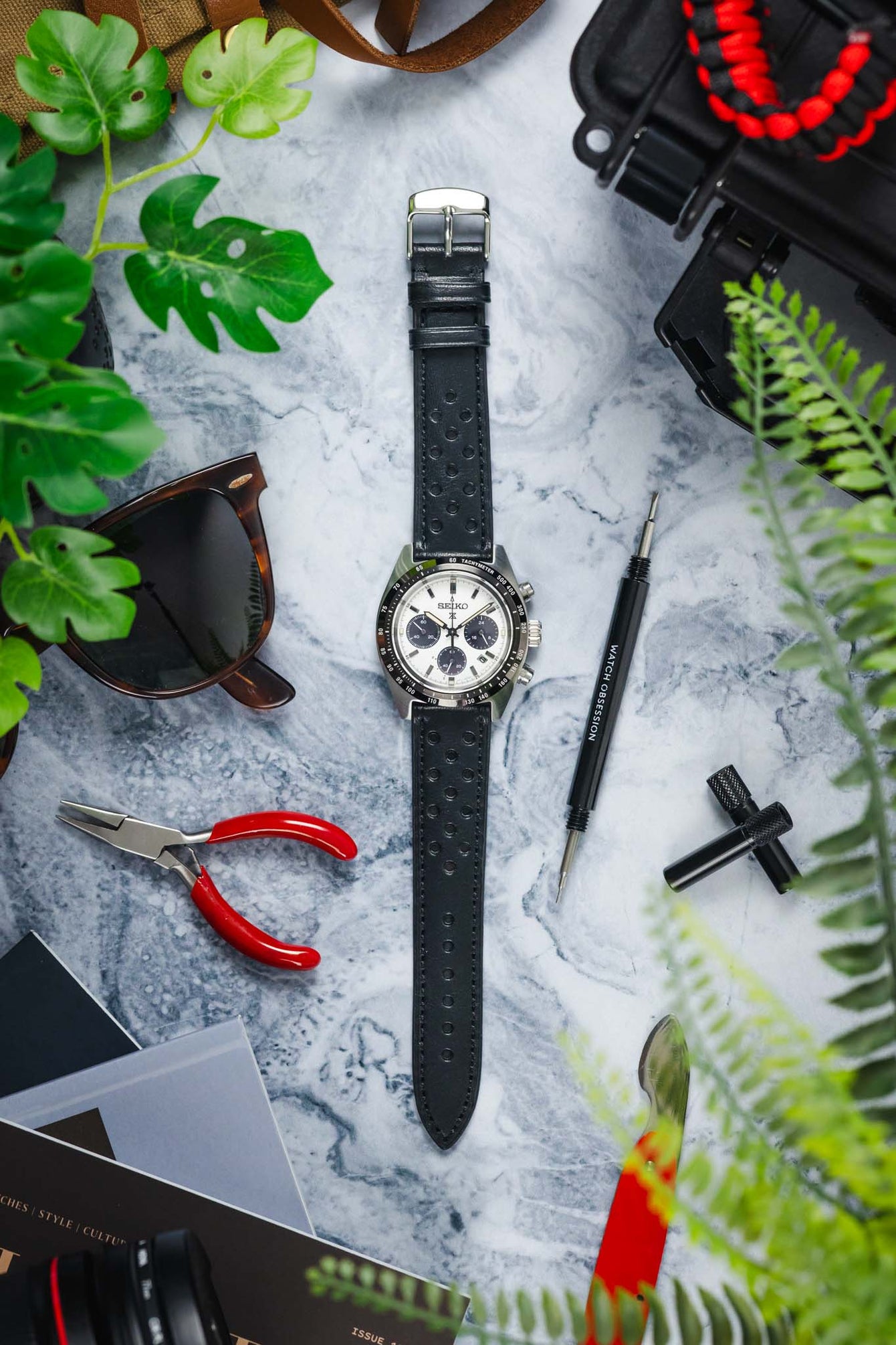 Flat lay of a Seiko chronograph wristwatch with a white dial and three dark subdials, mounted on a black perforated leather strap, arranged vertically on a marble surface. Surrounding items include a spring bar tool labeled “WATCH OBSESSION,” red-handled pliers, a black case, sunglasses, a magazine, and green plant leaves partially framing the scene.