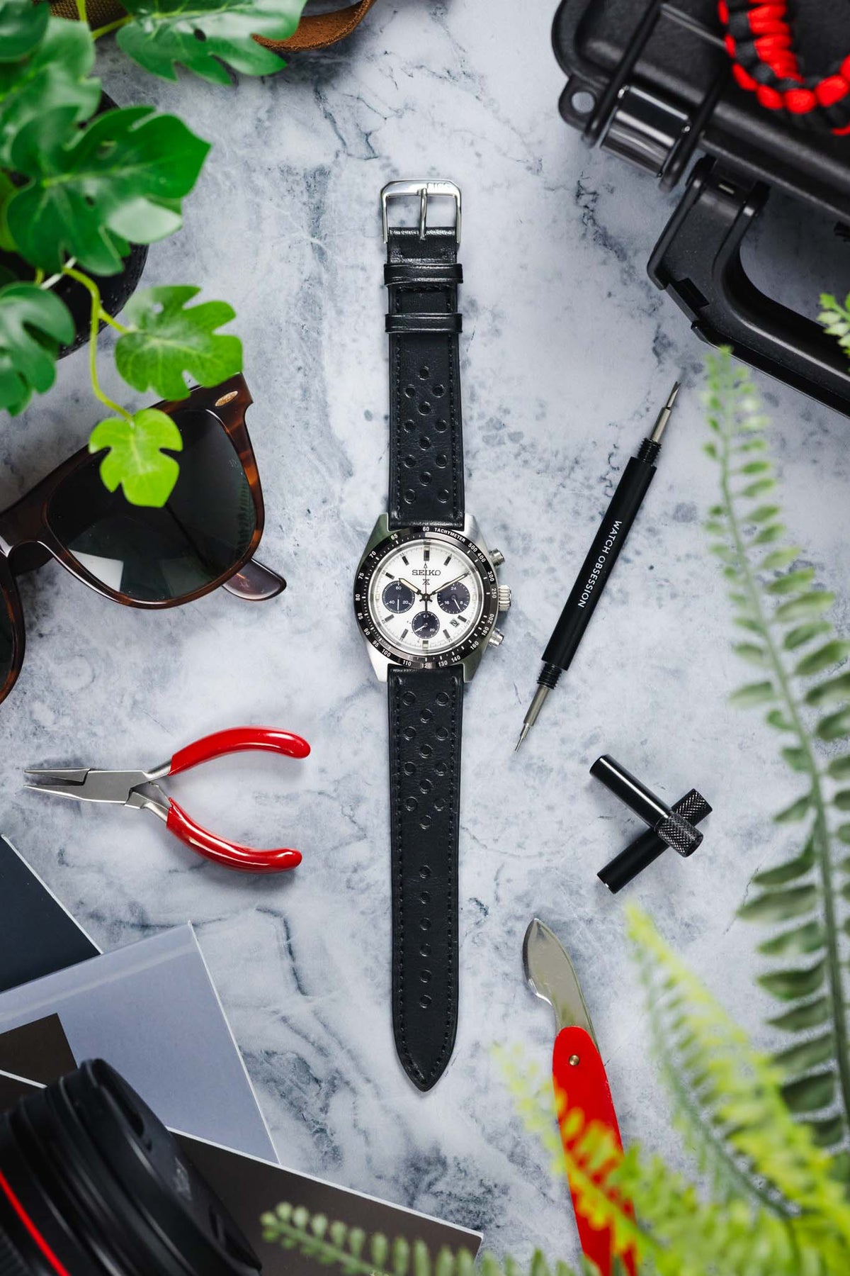 Seiko Speedtimer chronograph watch with a white panda dial, three black sub-dials, black tachymeter bezel, and a black perforated leather rally strap, displayed on a marble-effect surface alongside watchmaking tools, sunglasses, camera equipment, and greenery, highlighting the strap's classic motorsport-inspired design and premium handcrafted leather construction.