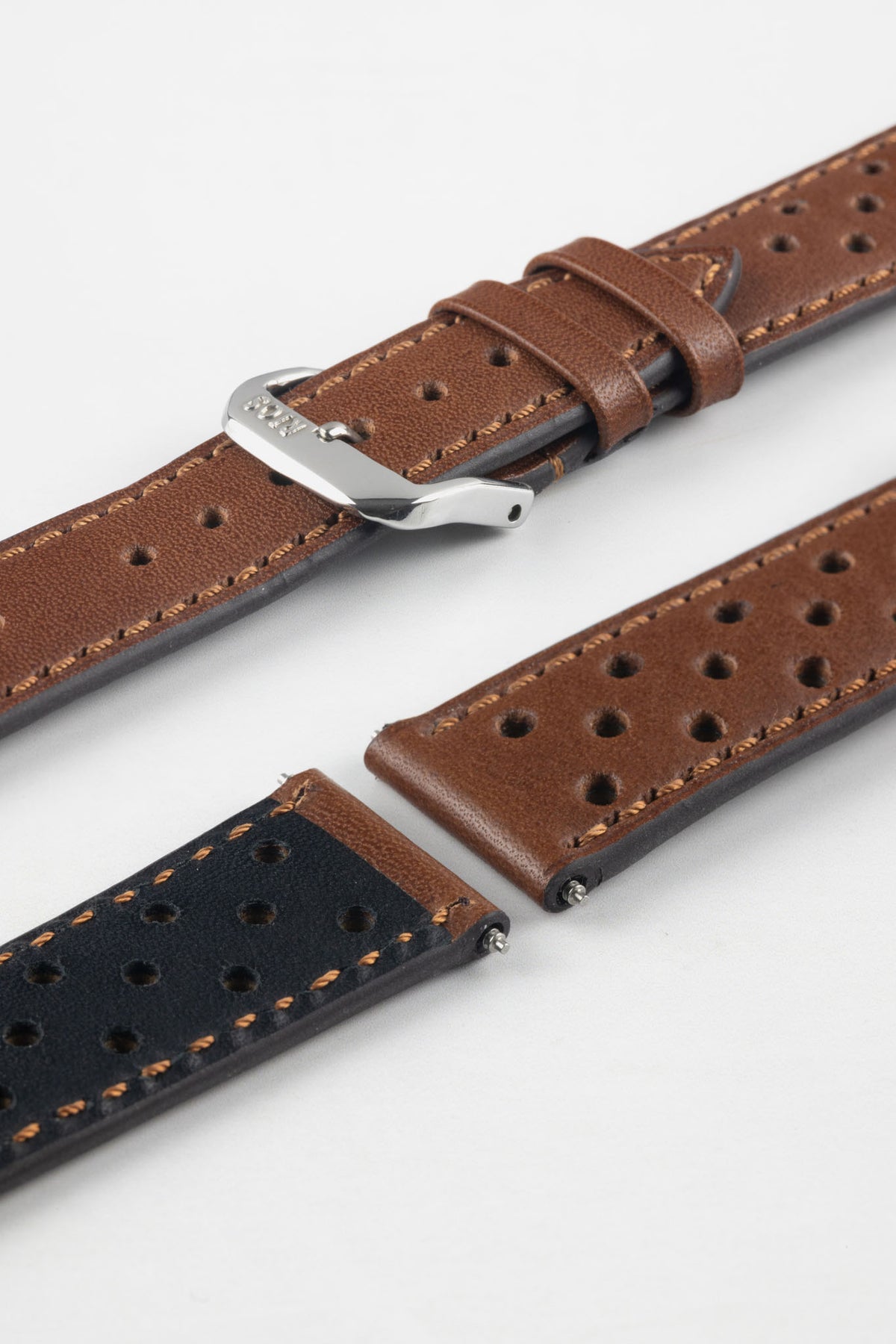 Close-up detail view of a cognac brown perforated leather rally watch strap displayed with one strap section detached, featuring matching brown stitching, multiple motorsport-inspired perforations, dual keeper loops, a polished stainless steel RIOS buckle, quick-release spring bars, and a black leather lining. Displayed on a light grey background to highlight the premium handcrafted construction, contrasting underside, and vintage racing-inspired design.