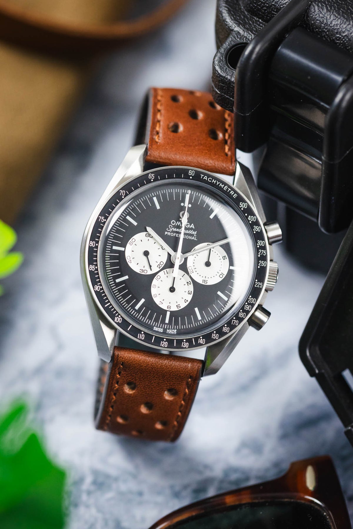 Close-up of an Omega Speedmaster Professional chronograph watch featuring a black dial with three white chronograph sub-dials, black tachymeter bezel, and polished stainless steel case, fitted with a cognac brown perforated leather rally strap with matching brown stitching. Displayed beside a black travel case on a marble-effect surface, the image highlights the strap's vintage motorsport-inspired design, premium handcrafted leather construction, and rich warm-toned finish.